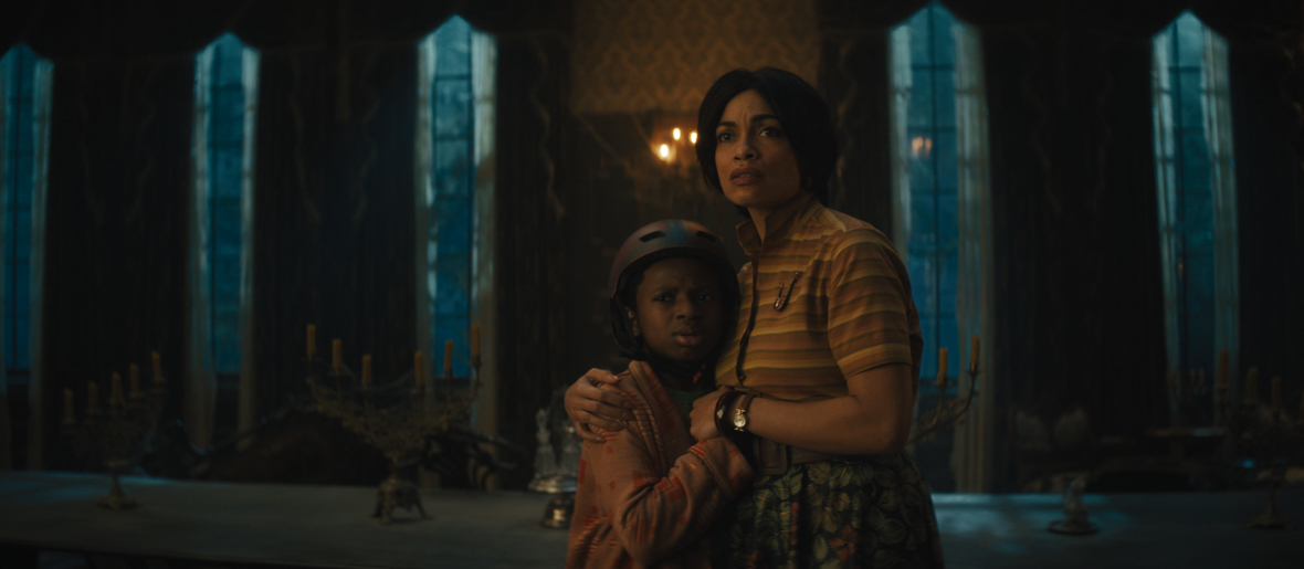 In an image from Disney’s Haunted Mansion (2023), Travis (Chase Dillon) and Gabbie (Rosario Dawson) clutch each other in the darkly lit dining room of the New Orleans mansion they’ve recently moved into—looking with concern at something beyond the camera.