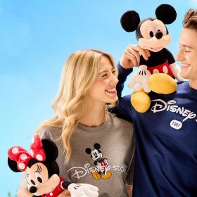 Two people holding Mickey and Minnie Mouse plush toys, both wearing Disney Store-themed apparel. One person wears a gray T-shirt with a classic Mickey design and 'Disney Store' text, while the other sports a navy long-sleeve shirt with 'Disney Store' and '1987' printed on it. The photo highlights nostalgic Disney merchandise and apparel, evoking a sense of playful celebration and brand heritage.