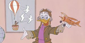 Professor Ludwig Von Drake, an animated duck character, wears an old-fashioned aviator costume of tan shirt, brown jacket with gold trim and buttons, lavender scarf, and, on the top of his head, goggles. He appears to be speaking, with his right hand extended and his left hand holding a model of a biplane with a spinning propellor. Ludwig is looking at the airplane model. A globe is partially visible on the right and there’s a framed image of a hot-air balloon with a solo pilot in an attached basket on the wall behind him.