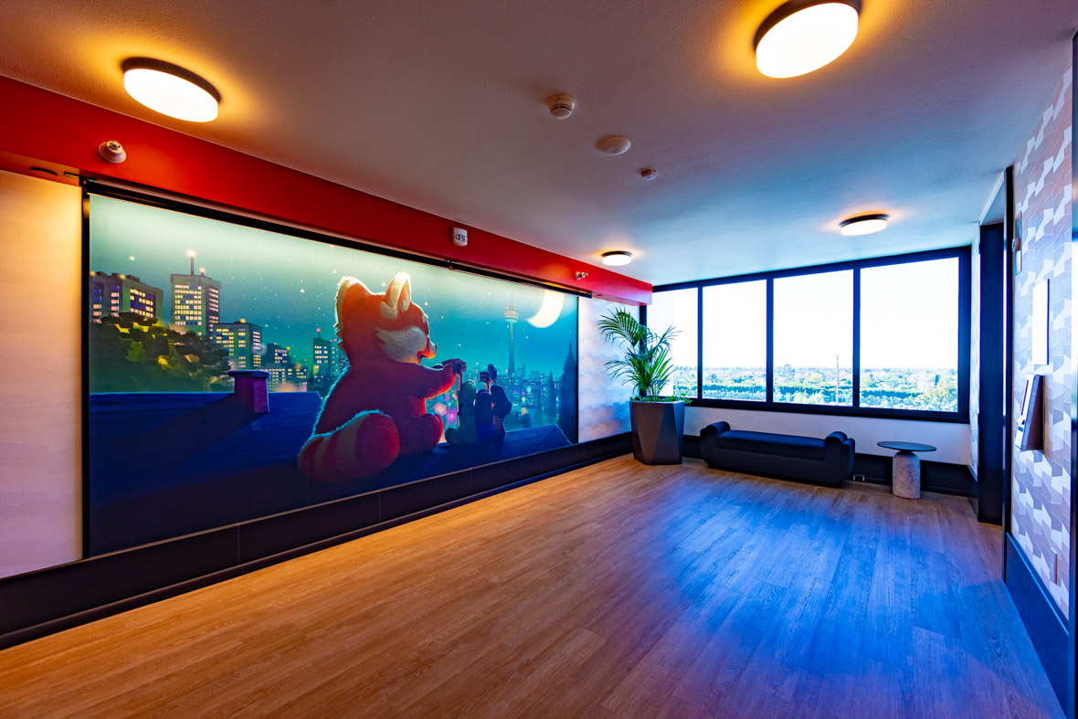 A hallway in Pixar Place Hotel, depicting a mural of panda Mei and her friends from Turning Red sitting on top of a building and looking out over the skyline. The mural is accented by a strip of red above the image. A wall perpendicular and to the right of the mural features actual windows looking out to a line of trees and sky.