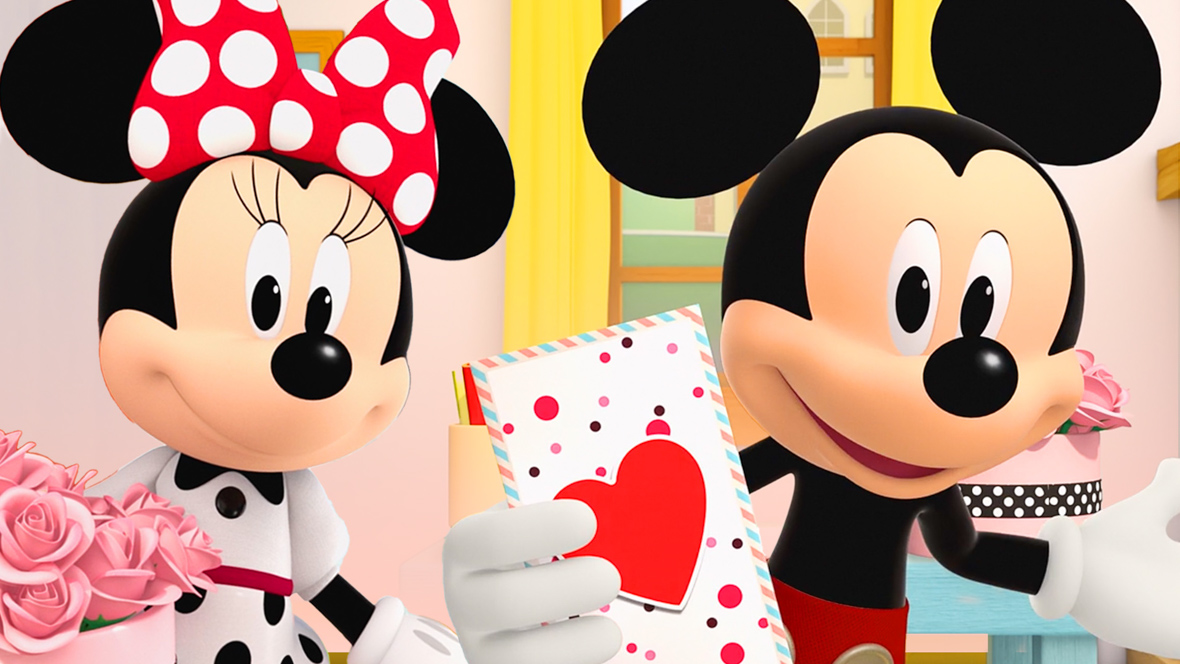 Mickey (right) and Minnie (left) stand facing the camera, displaying Valentine's Day gifts. Mickey holds up a card adorned with a heart, while Minnie, dressed in a polka dot dress and bow, holds a bouquet of pink roses.
