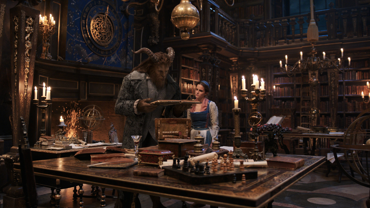 Belle (left), portrayed by Emma Watson, and The Beast (right), portrayed by Dan Stevens, are in The Beast’s library, a grand room with a high vaulted ceiling, intricately carved walls and ceiling, adorned with gold accents. Belle wears a white, long-sleeved underdress, layered with a blue, short-sleeved gown, complemented by a white apron and a blue headband. The Beast wears a black coat with gray detailing, a gray shirt, and black pants. He is holding a book that he and Belle are observing.  