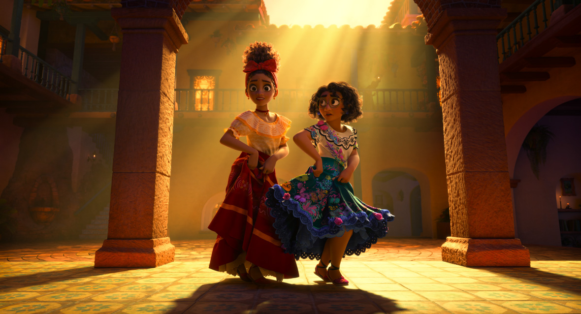 Mirabel and Dolores joyfully dance in a brick, stone, and ceramic open-space patio. Mirabel wears round green glasses and a vibrant blue and white dress adorned with embroidered detailing, fringe, tassel trims, and butterfly appliques. Dolores has a red bow adorning the front of her head and wears a charming red, white, and yellow dress featuring lace around the collar, puff sleeves, and striped detailing on the skirt.