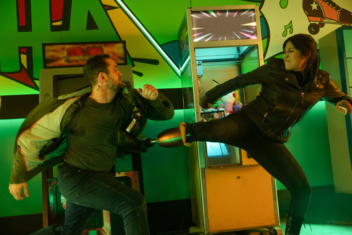In a scene from Marvel Studios’ Echo, one of Wilson Fisk’s thugs (on the left) crouches with his right foot forward. Maya Lopez, played by Alaqua Cox, uses her prosthetic leg to kick the man’s ribcage. They are fighting near an old arcade game inside an outdated bowling alley.
