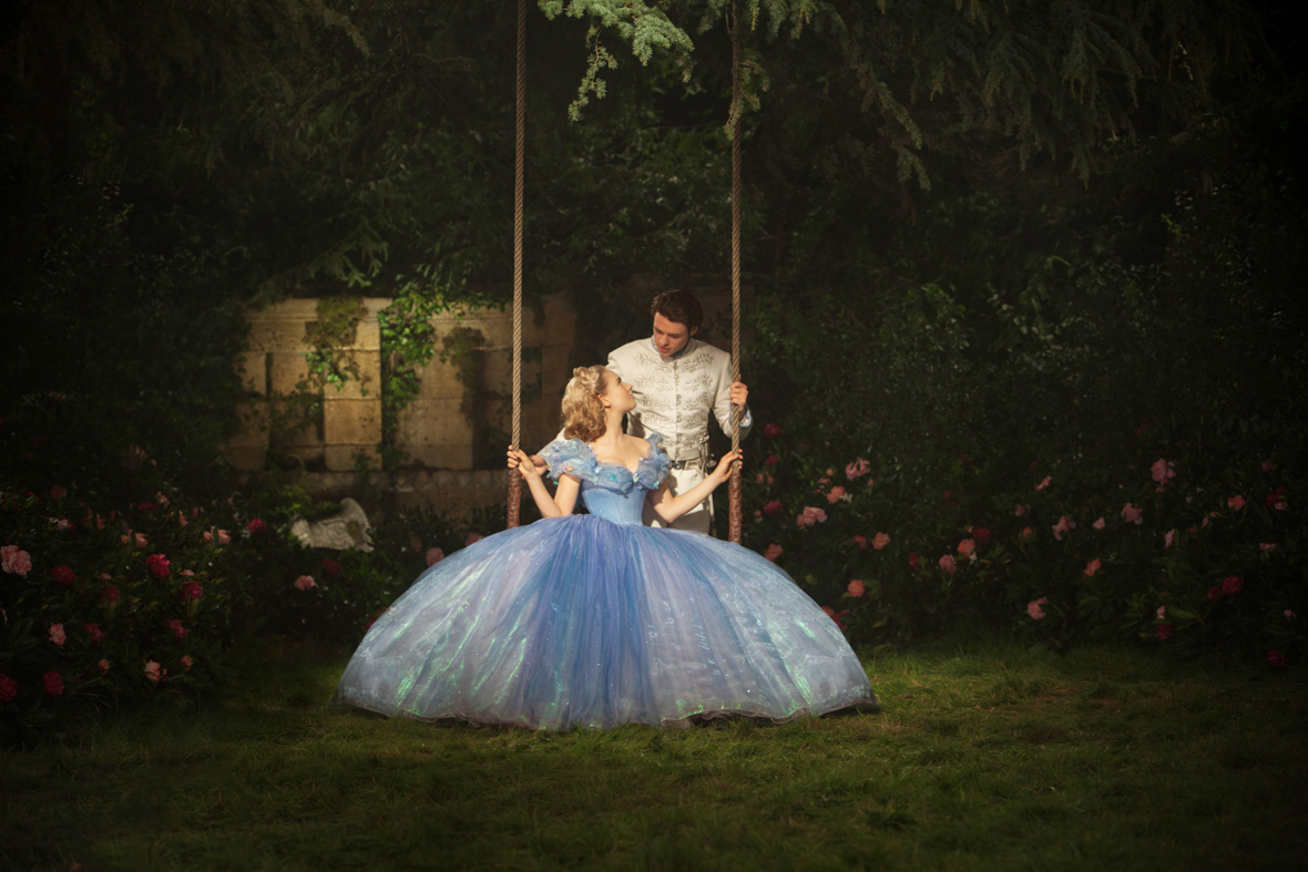 Cinderella (left), played by Lily James, sits gracefully on a low swing, wearing a voluminous, off-the-shoulder blue gown. Prince Kit (right), played by Richard Madden, stands behind her, dressed in a regal white jacket and pants. Their eyes lock in a tender gaze amidst a picturesque garden adorned with trees and red and pink roses.