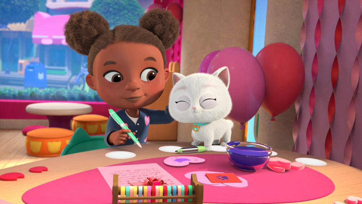 Bitsy (left) sits on a wooden table while being pet by Girl (right), who holds a green pen and is in the midst of writing a Valentine’s Day card. The Girl wears a navy blue cardigan with small pink hearts.