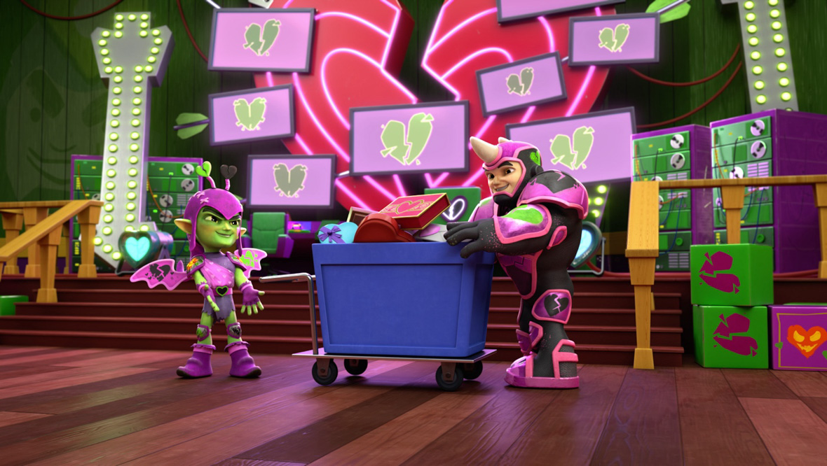 Gobby (left) and Rhino (right) are dressed in their Super Hero costumes and are placing all the Valentine’s Day gifts they have stolen into a blue cart. They are in a room with a wooden floor, green neon lights, and TV screens that feature broken green hearts.