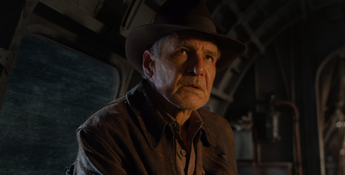 Indiana Jones, played by Harrison Ford, is in a train, looking above with a worried and perplexed expression. Along with his fedora, Indiana Jones dons a rugged leather jacket, and a khaki shirt. 