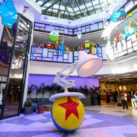 The atrium of Pixar Place Hotel, featuring a sculpture of the Pixar Lamp and the Pixar Ball in the center of the room. Hanging from the glass ceiling are mobiles depicting iconic Pixar characters in simple shapes and colors. Around the perimeter of the first two floors of the atrium, Disney cast members are celebrating the hotel’s opening.