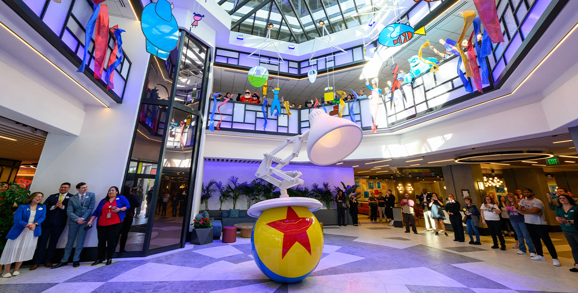 The atrium of Pixar Place Hotel, featuring a sculpture of the Pixar Lamp and the Pixar Ball in the center of the room. Hanging from the glass ceiling are mobiles depicting iconic Pixar characters in simple shapes and colors. Around the perimeter of the first two floors of the atrium, Disney cast members are celebrating the hotel’s opening.