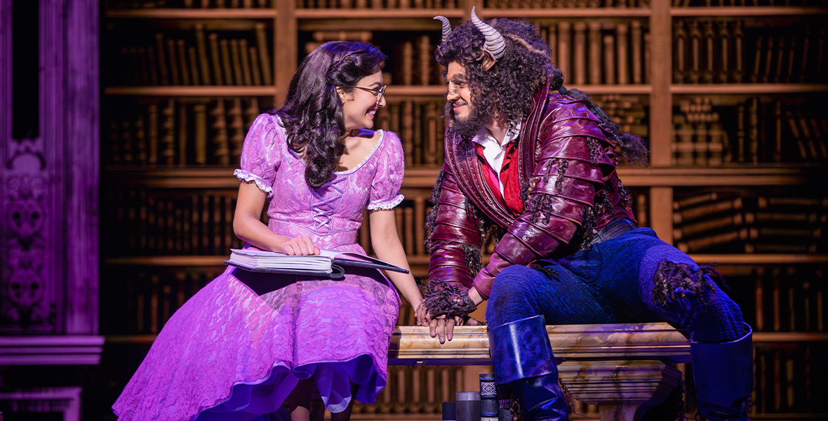 Belle (Shubshri Kandiah) and Beast (Brendan Xavier) sit on an upholstered bench in Beast’s library, each turned toward the other, in a scene from the current Australian production of the stage musical Beauty and the Beast.