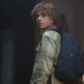 Annabeth, played by Leah Jeffries, Percy, played by Walker Scobell, and Grover, played by Aryan Simhadri, glance backward looking confused and fearful. Annabeth is dressed in a purple corduroy jacket, Percy sports a green flannel shirt with a blue backpack, and Grover dons a blue waterproof jacket.