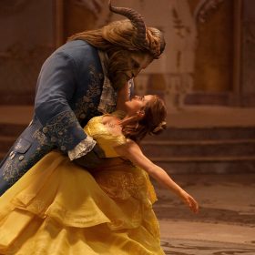 Belle (right), played by Emma Watson, is dancing with The Beast (left), played by Dan Stevens. Belle is wearing a voluminous, off-the-shoulder, yellow ballgown, and The Beast is wearing a navy blue ballroom tailcoat trimmed with gold. The Beast holds Belle from her waist, as she leans backward, holding The Beast by his neck. They gaze into each other’s eyes.