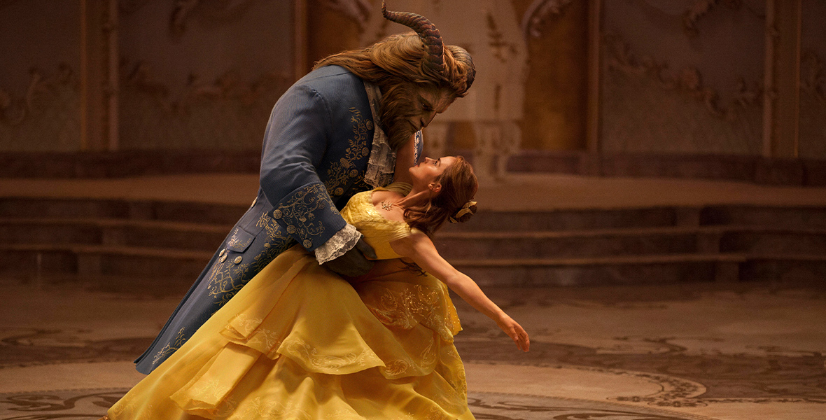 Belle (right), played by Emma Watson, is dancing with The Beast (left), played by Dan Stevens. Belle is wearing a voluminous, off-the-shoulder, yellow ballgown, and The Beast is wearing a navy blue ballroom tailcoat trimmed with gold. The Beast holds Belle from her waist, as she leans backward, holding The Beast by his neck. They gaze into each other’s eyes.
