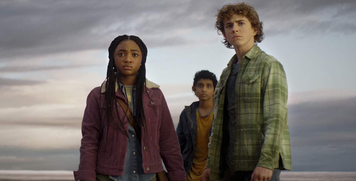 In an image from Disney+’s Percy Jackson and the Olympians, from left to right, Annabeth (Leah Sava Jeffries), Grover (Aryan Simhadri), and Percy (Walker Scobell) are standing on a beach looking at something off camera to the left. The skies behind them are stormy. Annabeth is wearing a purple jacket and green pants; Grover is wearing a blue long-sleeved shirt over a yellow t-shirt and khaki pants; and Percy is wearing a green plaid long-sleeved shirt and jeans.