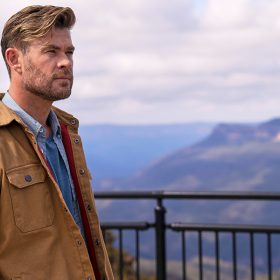 three-fourths toward the camera and contemplates a vista to the right. He wears a tan jacket over a light blue button-up shirt. A blue sky and mountains are in the backdrop.