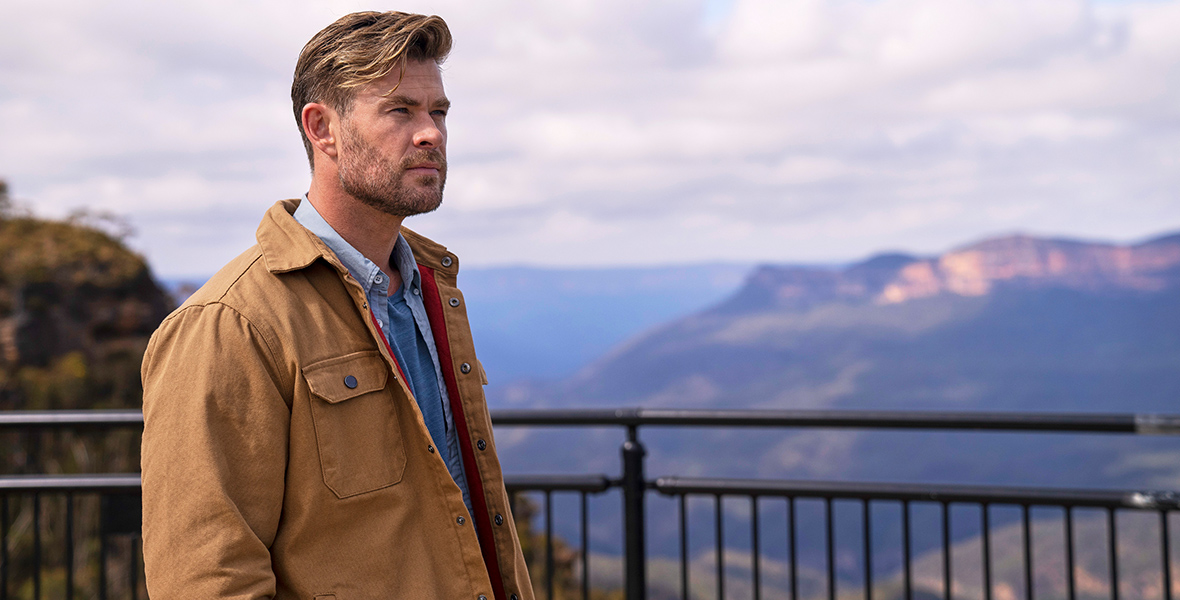 three-fourths toward the camera and contemplates a vista to the right. He wears a tan jacket over a light blue button-up shirt. A blue sky and mountains are in the backdrop.