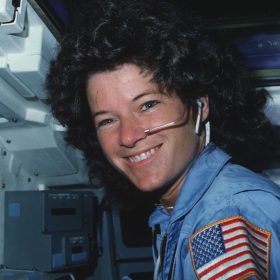 Mission Specialist Sally Ride as seen in a photo from June 24, 1983, as part of promotional material for the new documentary Sally. She is sitting inside a spacecraft, in a blue uniform with an American flag patch on the upper arm. She smiles broadly toward the camera, with a face mic visible by her mouth.