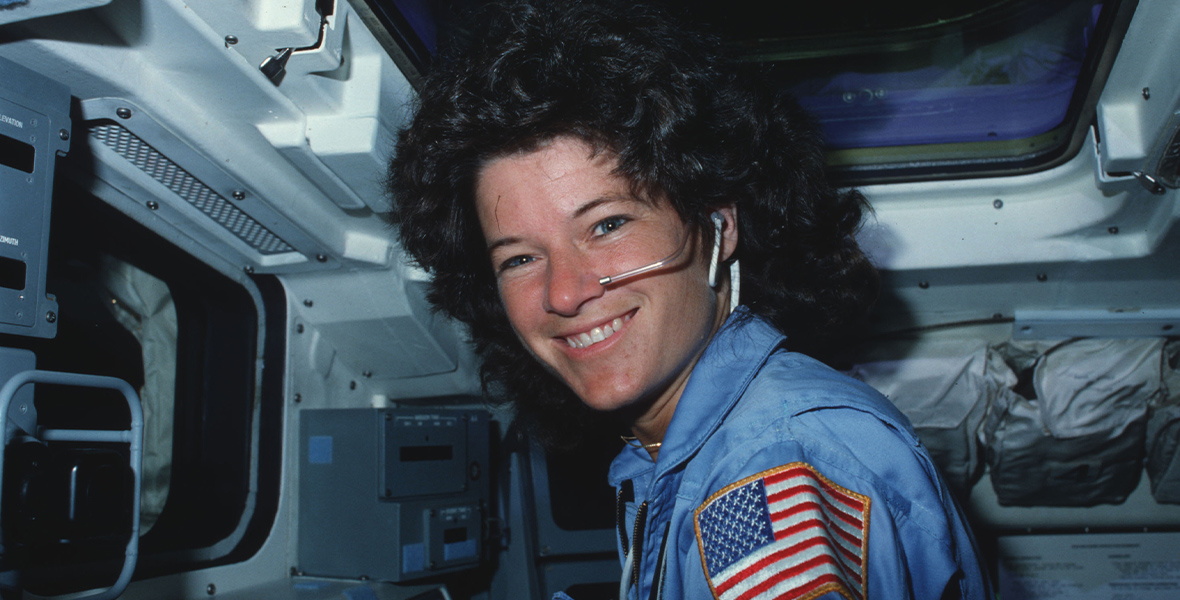 Mission Specialist Sally Ride as seen in a photo from June 24, 1983, as part of promotional material for the new documentary Sally. She is sitting inside a spacecraft, in a blue uniform with an American flag patch on the upper arm. She smiles broadly toward the camera, with a face mic visible by her mouth.
