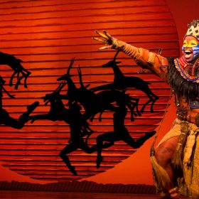 Actor Mukelisiwe Goba performs on stage as Rafiki in Disney Theatricals’ The Lion King, with a large orange sun and silhouette of antelopes in the background. Goba is singing with power and holding their hand to the sky.