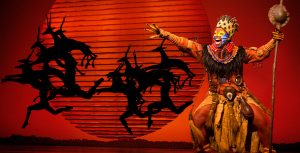 Actor Mukelisiwe Goba performs on stage as Rafiki in Disney Theatricals’ The Lion King, with a large orange sun and silhouette of antelopes in the background. Goba is singing with power and holding their hand to the sky.