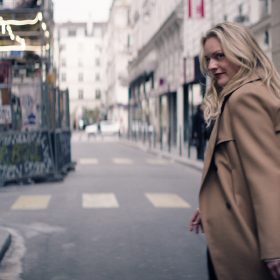 Elizabeth Moss, in the role of Imogen Salter, is seen in a promotional still for the FX spy thriller The Veil. She stands at the far right in profile with her face turned back toward the camera with a sly smile of her face. Her long blond hair is draped over her back. She wears a tan trench coat and is standing in an alley in an urban environment.