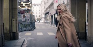 Elizabeth Moss, in the role of Imogen Salter, is seen in a promotional still for the FX spy thriller The Veil. She stands at the far right in profile with her face turned back toward the camera with a sly smile of her face. Her long blond hair is draped over her back. She wears a tan trench coat and is standing in an alley in an urban environment.