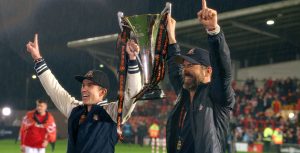 On the field of a sports stadium, Rob McElhenny, on the left, and Ryan Reynolds, owners of the professional football league soccer team from Wrexham, U.K., hoist a trophy between them with their index fingers raised in the gesture for No. 1 on a soccer field as rain falls. They are smiling broadly and are outfitted in Wrexham-logoed gear.