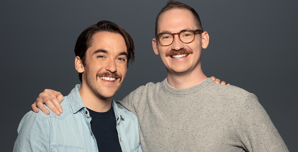 Chris Houghton (left) wears a denim jacket over a black T-shirt. Shane Houghton (right) wears glasses and a gray sweater. Both brothers have mustaches and are smiling.