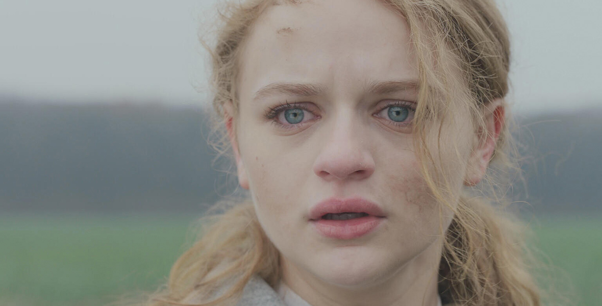 In a promotional image for Hulu’s We Were the Lucky Ones, Joey King, who plays Halina, is seen in close-up as she looks out at something in the distance. Her blue eyes are watery and it looks like she has been crying. Her long blond hair is tied back with a long wisp falling over her face. She stands in a hazy natural setting.