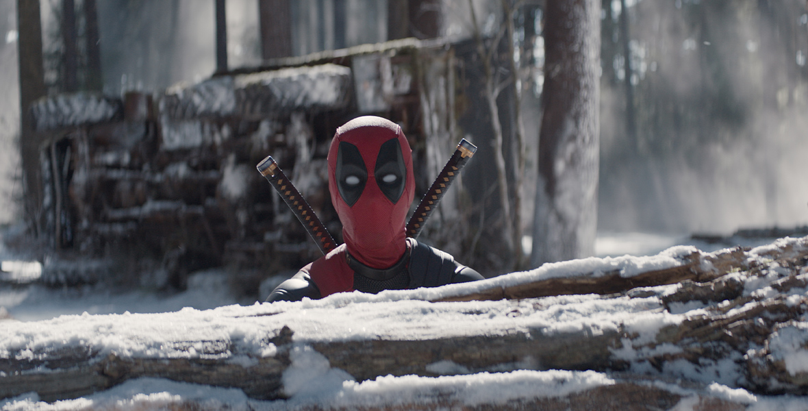 Deadpool, masked here in a red and black costume, peeks his head over a snowy tree trunk. He is outside in a snowy forest, looking directly into the camera.