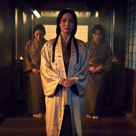 wears a white silk robe with black details over a brown kimono with orange patterns. Her long black hair falls forward in two strands, and she looks to her right. Two women beside her wear brown kimonos with patterns, their heads slightly bent forward in respect. This scene unfolds within a candlelit, Japanese-style hallway characterized by wooden flooring and traditional windows.