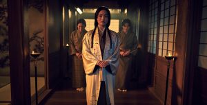 wears a white silk robe with black details over a brown kimono with orange patterns. Her long black hair falls forward in two strands, and she looks to her right. Two women beside her wear brown kimonos with patterns, their heads slightly bent forward in respect. This scene unfolds within a candlelit, Japanese-style hallway characterized by wooden flooring and traditional windows.