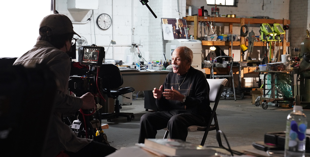 Former test pilot Ed Dwight sits down for an interview about National Geographic’s The Space Race. He is wearing a black crewneck sweater and black pants. While speaking to a filming camera, a microphone hovers above him. The background features tools hanging from wooden shelves.