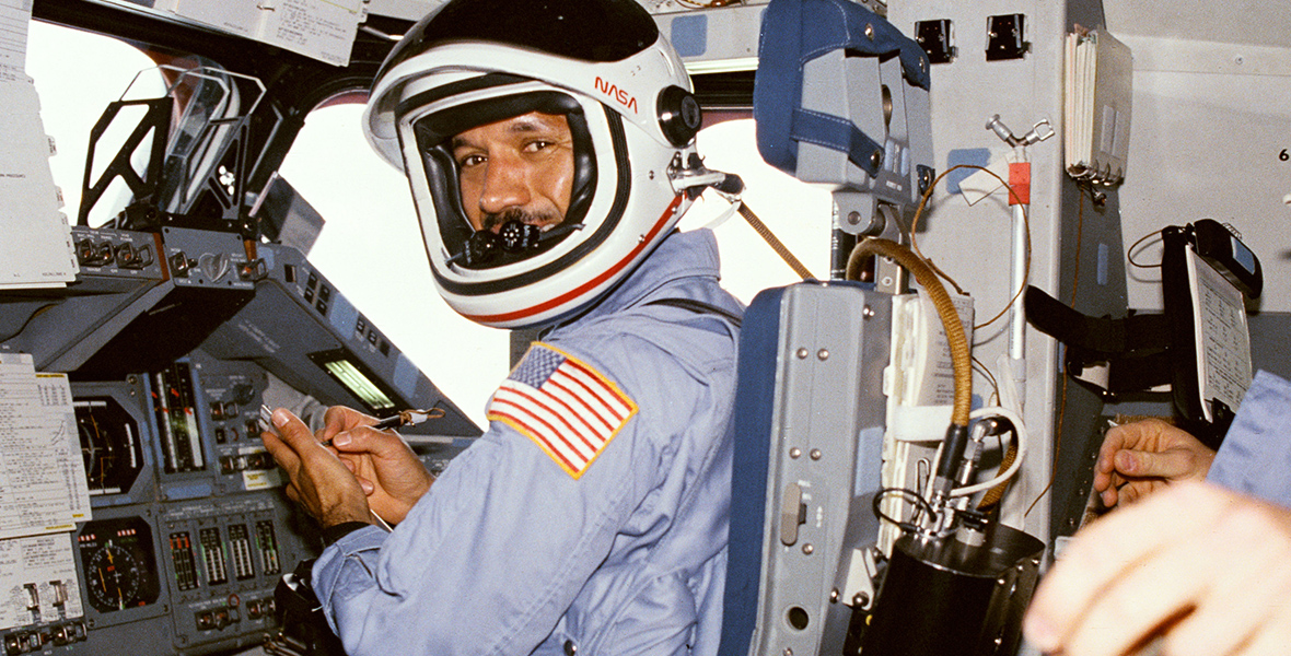 In an image from National Geographic Documentary Films’ The Space Race, astronaut Charlie Bolden is seen on the shuttle Columbia. He is wearing a blue jumpsuit with the American flag sown on his left shoulder and a white helmet that reads “NASA” with red letters. 