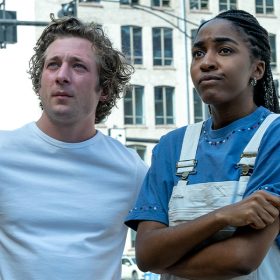 In a scene from FX’s The Bear, Carmen “Carmy” Berzatto, played by Jeremy Allen White, and Sydney Adamu, played by Ayo Edebiri, stand on Chicago a street corner. Carmy is wearing a white T-shirt, which exposes several tattoos on his arms, and has both his hands on his waist. Sydney is wearing a blue shirt and white overalls and crosses her arms.