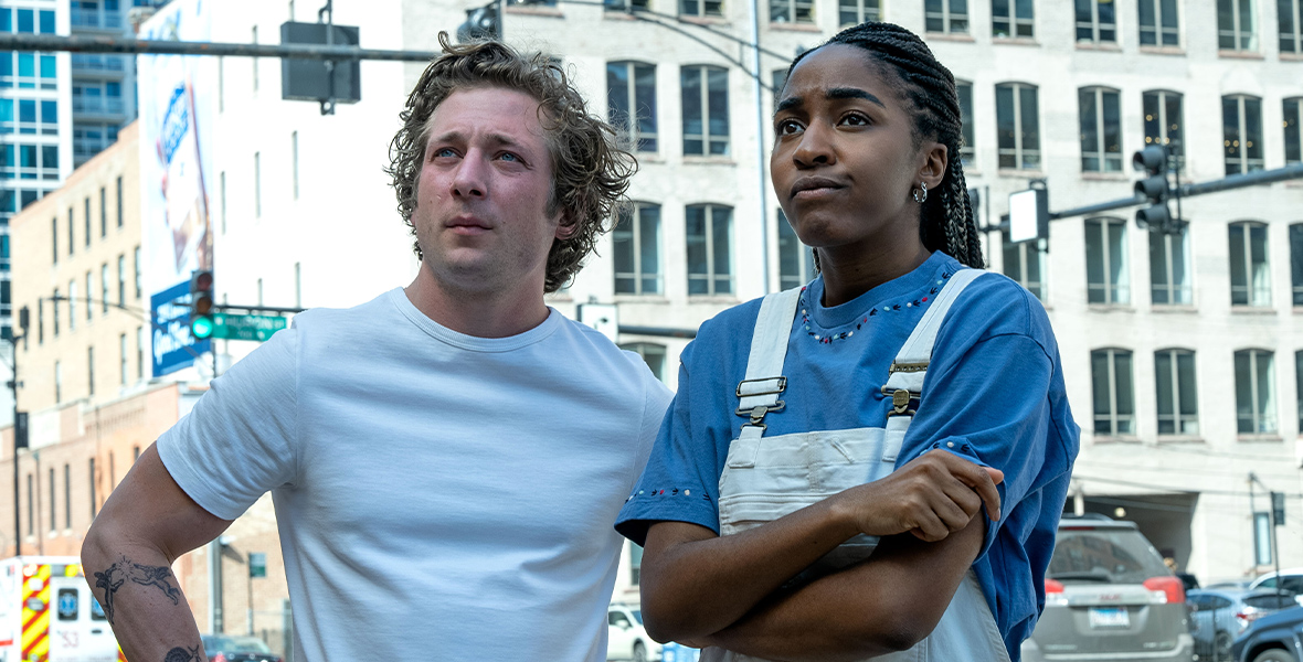 In a scene from FX’s The Bear, Carmen “Carmy” Berzatto, played by Jeremy Allen White, and Sydney Adamu, played by Ayo Edebiri, stand on Chicago a street corner. Carmy is wearing a white T-shirt, which exposes several tattoos on his arms, and has both his hands on his waist. Sydney is wearing a blue shirt and white overalls and crosses her arms.