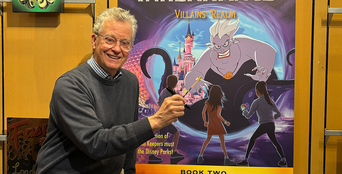 New York Times best-selling author Ridley Pearson stands to the left, turned three-quarters toward the camera in front of a large display of the book cover of Volume 2 of Kingdom Keepers Inheritance: Villains’ Realm. He wears clear-rimmed eyeglasses and is smiling and is raising his left hand so it appears like it is holding a wand topped by a star that is seen on the book cover. He is wearing a checkered-collared shirt beneath a gray sweater. The book jacket features a large image of Disney Villain Ursula with Cinderella’s castle from Walt Disney World and famed EPCOT sphere visible on the left at night. The book jacket text reads: “New York Times best-selling author Ridley Pearson Kingdom Keepers Inheritance.” A smaller piece of art showing Maleficent at the center is hung to the left of the book jacket on the back wall.