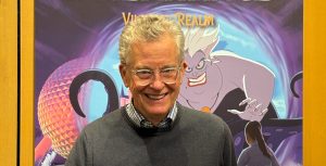 New York Times best-selling author Ridley Pearson stands in front of a large display of the book cover of Volume 2 of Kingdom Keepers Inheritance: Villains’ Realm. He wears clear-rimmed eyeglasses and is smiling and holding a pen in his left hand. He is wearing a checkered-collared shirt beneath a gray sweater. The book jacket features a large image of Disney Villain Ursula with the famed EPCOT sphere visible on the left at night. The book jacket text reads: “New York Times best-selling author Ridley Pearson Kingdom Keepers Inheritance.”