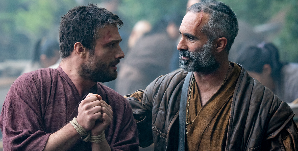 In a scene from FX's Shōgun, John Blackthorne, played by Cosmo Jarvis, has rope handcuffs around his wrist. He looks to his right at Rodrigues, played by Nestor Carbonell.