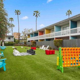 Outdoor lawn area with colorful Adirondack chairs, oversized games such as Connect Four and chess, surrounded by vibrant two-story buildings featuring colorful accents and balconies. Palm trees and a clear blue sky add to the lively atmosphere