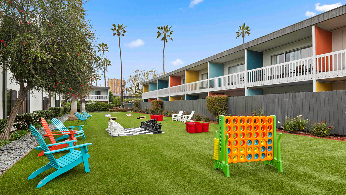 Outdoor lawn area with colorful Adirondack chairs, oversized games such as Connect Four and chess, surrounded by vibrant two-story buildings featuring colorful accents and balconies. Palm trees and a clear blue sky add to the lively atmosphere