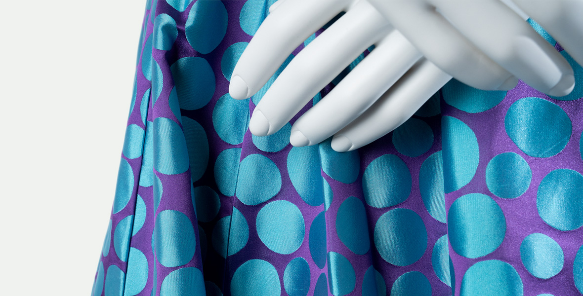 A close-up of a section of a costume designed by Sandy Powell for Anastasia (Holliday Grainger) in Cinderella (2015) shows silky purple fabric with big blue polka dots, slightly obscured by a mannequin’s white hand.