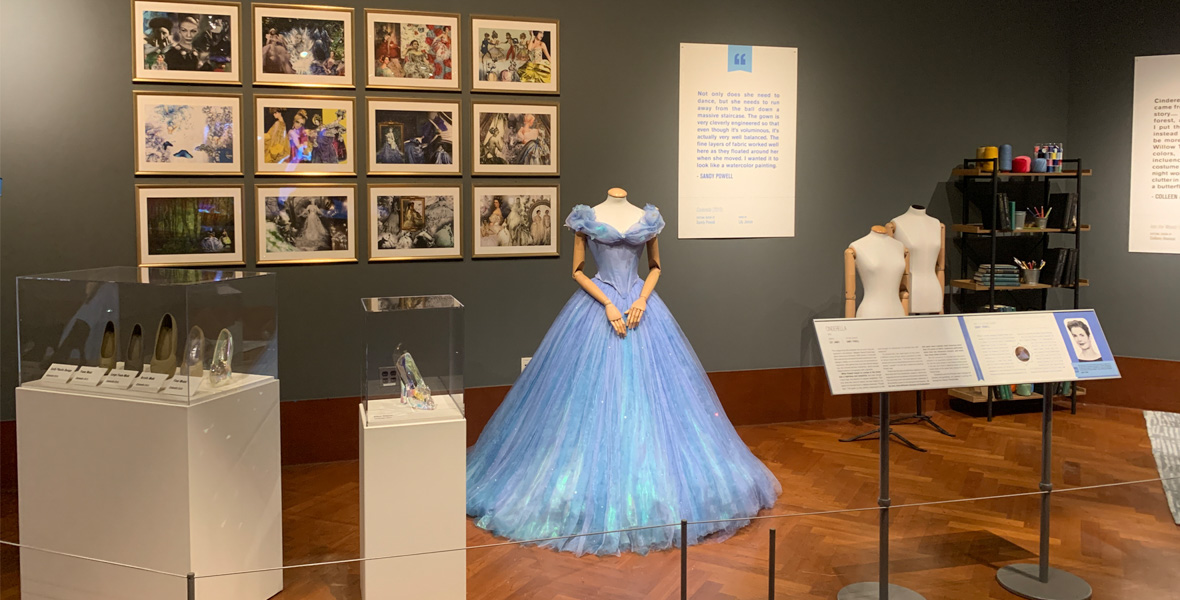 The “Cinderella’s Workshop” gallery is seen as presented in Heroes & Villains: The Art of the Disney Costume at the Henry Ford Museum of American Innovation in 2022. At the center is Cinderella’s blue ball gown, with a display of glass slippers and other footwear in glass cases to the left, and a display of a dozen colorful hand-drawn images, seen here too small to make out the details, on the wall behind the dress.