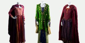 Headless mannequins display three costumes designed by Salvador P&eacute;rez Jr. for the Sanderson sisters (played by Bette Midler, Sarah Jessica Parker, and Kathy Najimy) in Hocus Pocus 2 (2022). All three are floor-length gowns with capes. The dresses on the right and left are crimson, while the middle dress is green.
