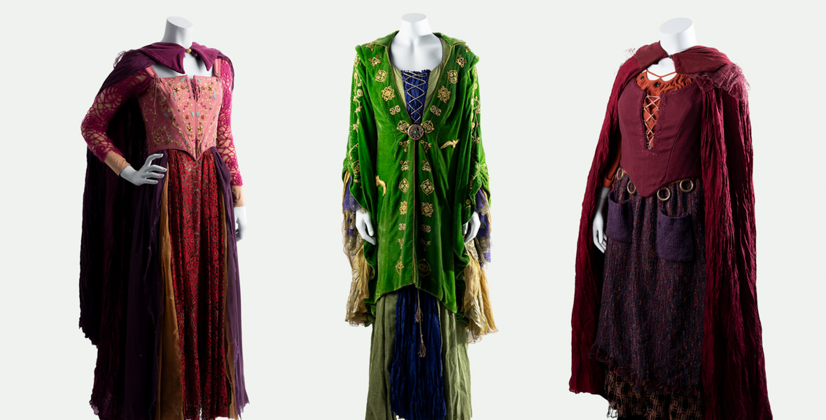 Headless mannequins display three costumes designed by Salvador Pérez Jr. for the Sanderson sisters (played by Bette Midler, Sarah Jessica Parker, and Kathy Najimy) in Hocus Pocus 2 (2022). All three are floor-length gowns with capes. The dresses on the right and left are crimson, while the middle dress is green.