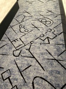 A gray rug in a hallway of Pixar Place Hotel. The rug features abstract line art in black of various Pixar characters, including Remy, Mater, and Edna Mode.