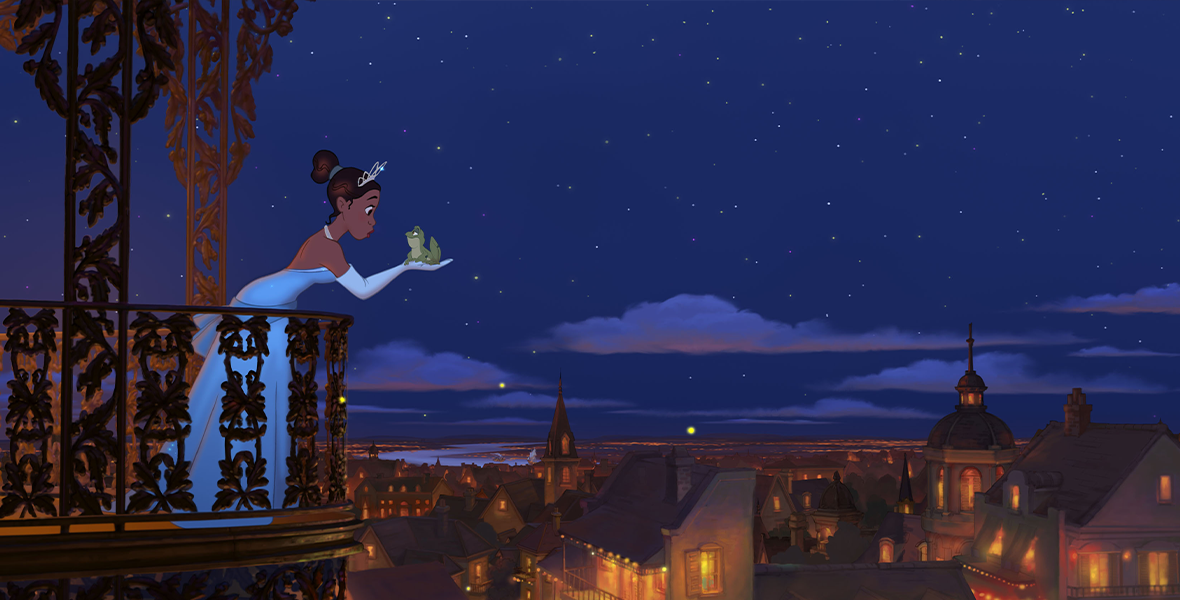 Still from The Princess and the Frog film, featuring Tiana holding Naveen in frog form on the balcony of Charlotte La Bouff’s Mansion. The New Orleans Skyline and a sky full of stars sit behind the pair.