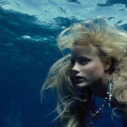 The mermaid Madison (played by Daryl Hannah) is seen from shoulders up below the surface of the blue water, her blond hair floating wildly around her head. She’s wearing an elaborate necklace studded with sealife-themed charms, beads, and coins.