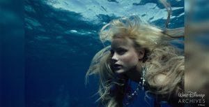 The mermaid Madison (played by Daryl Hannah) is seen from shoulders up below the surface of the blue water, her blond hair floating wildly around her head. She’s wearing an elaborate necklace studded with sealife-themed charms, beads, and coins.